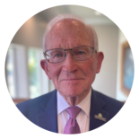 Portrait of an elderly man in a suit and tie, wearing glasses and a lapel pin, against a blurred background