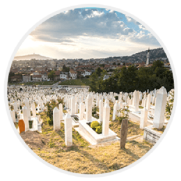 A hillside cemetery with rows of white gravestones under a cloudy sky, overlooking a city.