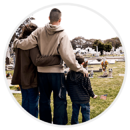 Three people standing together in a cemetery, facing a gravesite with a small American flag.