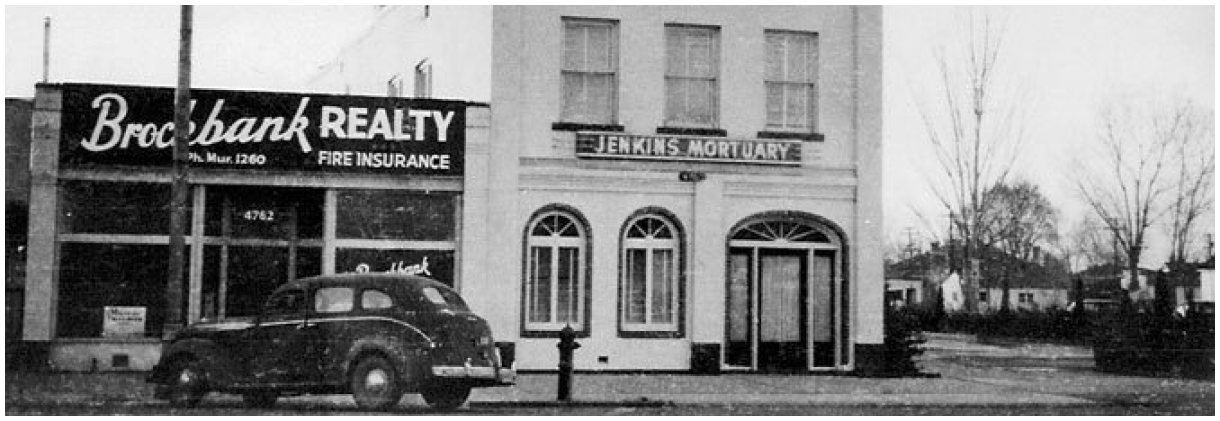 Black-and-white street scene with a realty storefront, parked truck, and adjacent building with arched windows