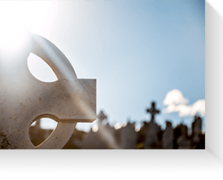 Metal cross sculpture silhouetted against a bright blue sky with clouds and sunlight.