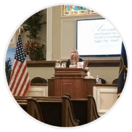 Speaker at a podium in a church-like hall, with an American flag and presentation screen behind her.