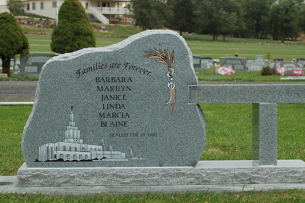 Gray memorial stone bench with engraved names and a cross in a grassy cemetery setting