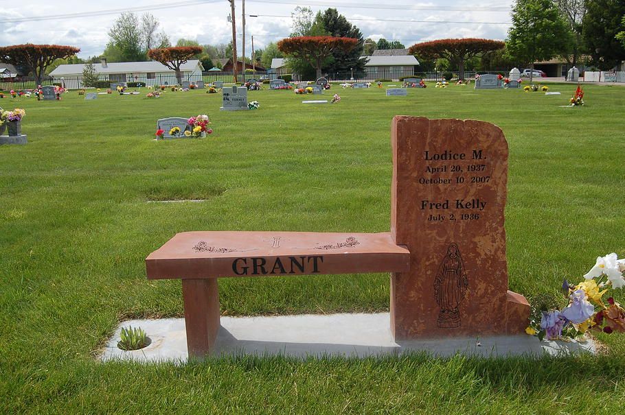 Gravestone and memorial bench in a green cemetery, with flowers and other graves in the background.