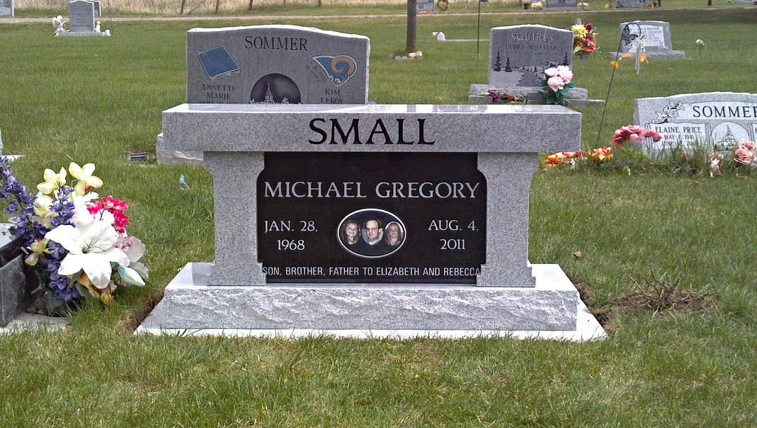 Gray cemetery headstone with “SMALL” engraved, surrounded by grass and flowers.