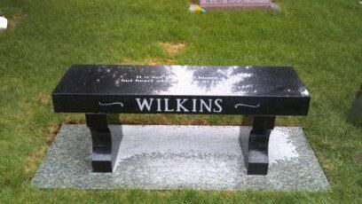 Black memorial bench with “WILKINS” on the back, set on a concrete pad in a grassy area.