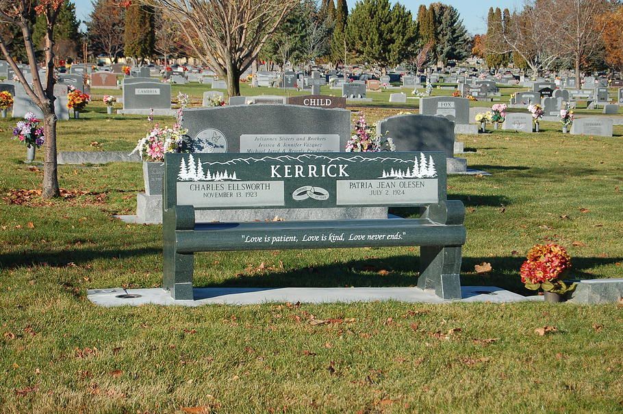 Gray memorial bench with “Kerrick” on it in a cemetery, with flowers nearby and graves in the background