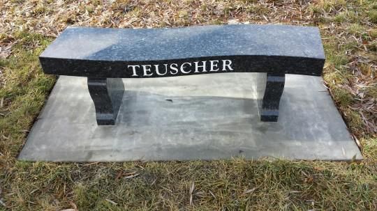Black stone memorial bench engraved “TEUSCHER” on a concrete base outdoors in grass