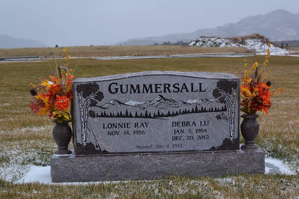 Gravestone in a snowy field with “GUMMERSALL” engraved, framed by flowers and mountains in the background
