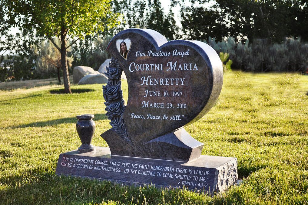 Heart-shaped black gravestone in a sunny cemetery, with white lettering and flowers on the base.