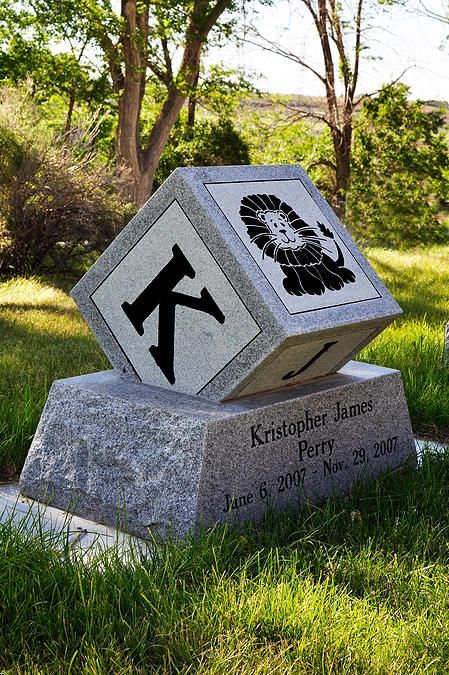 Gray cube sculpture on stone base in a grassy park, with black hiking and boot symbols on its faces