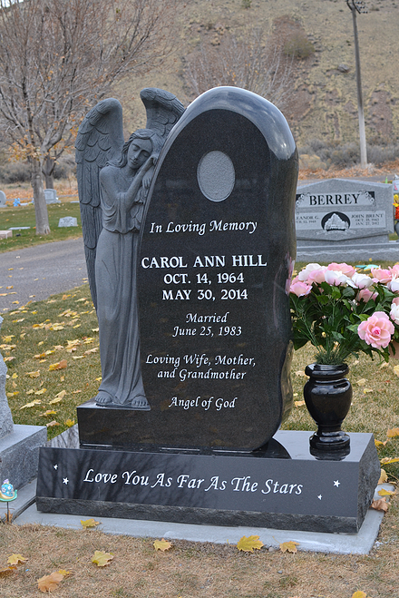 Black angel headstone with flowers in a cemetery; plaque reads “Love You To The Stars.”