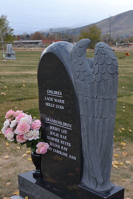 Black angel-wing gravestone with engraved text and pink flowers in a cemetery landscape.