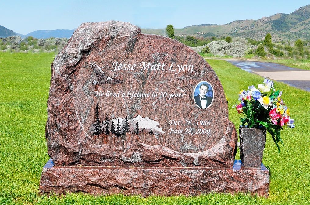 Engraved memorial stone in a green field, with flowers and a portrait plaque under a cloudy sky.