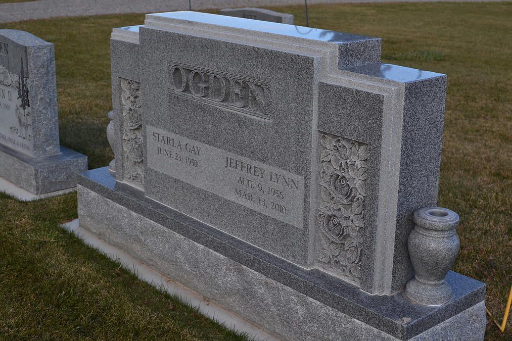 Gray granite cemetery tombstone with engraved plaques and decorative urns on a grassy plot