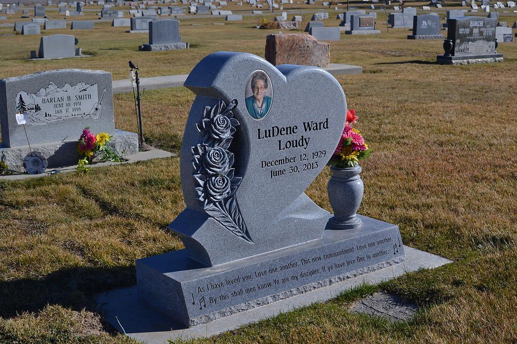 Heart-shaped gray gravestone with flowers in a cemetery of many markers under bright sunlight