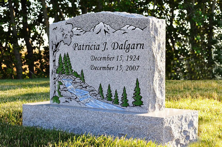 Gravestone with engraved deer, trees, and snow scene in a grassy cemetery at sunset