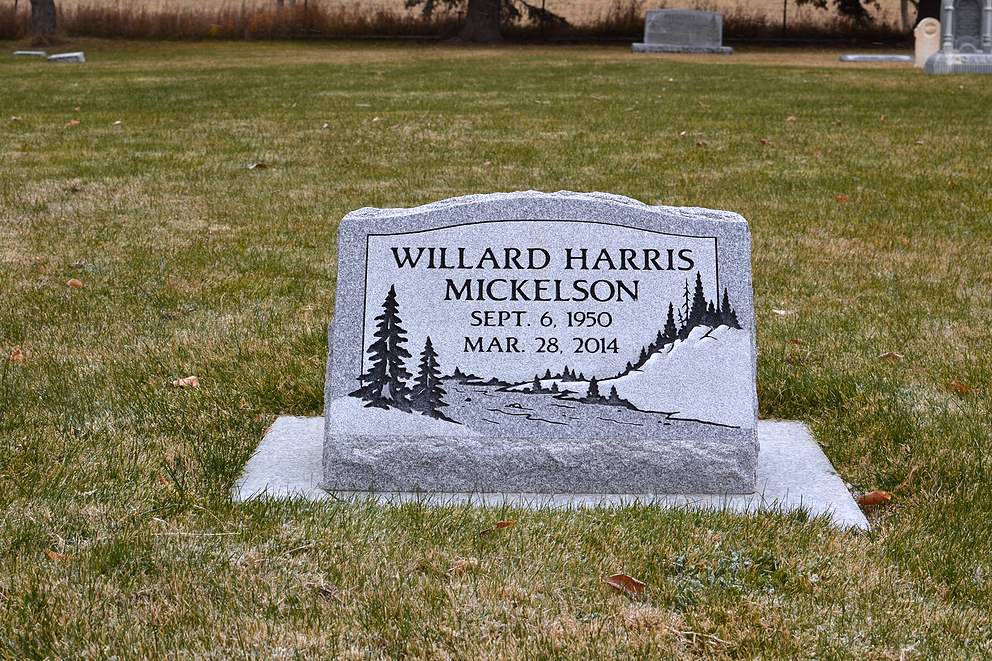 Gray gravestone in a grassy cemetery, engraved “Willard Harris Mickelson,” with trees carved on it.
