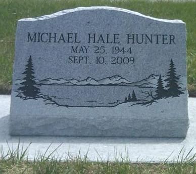 Gray granite gravestone with engraved name and dates, framed by pine tree and mountain scene