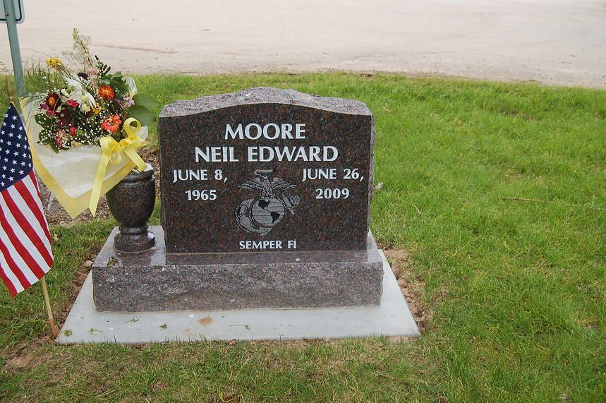 Gravestone in a grassy cemetery with flowers, a vase, and an American flag beside it.