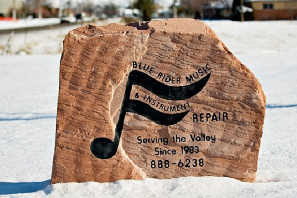 Wooden Bluebird Music repair sign in the snow, with a stylized black musical note and phone number.