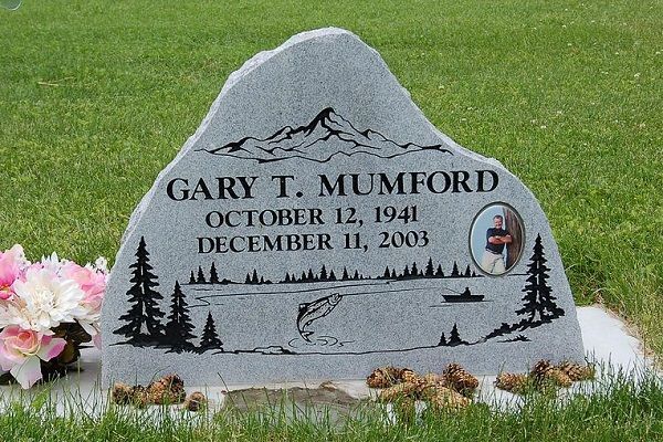 Gray memorial gravestone for Gary T. Mumford with flowers and engraved mountain and pine tree scene