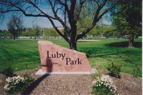 Stone sign reading “Luby Park” in a green park with trees and flower beds