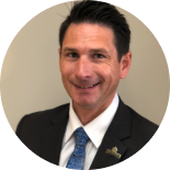 Professional headshot of a smiling person in a dark suit and blue tie against a beige background