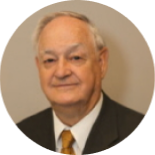 Portrait of an older man in a dark suit, white shirt, and gold tie, against a plain beige background