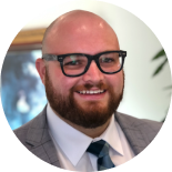 Smiling man with glasses and beard in a suit and tie, against a blurred indoor background