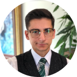 Portrait of a person in glasses and a green tie, wearing a suit, with a blurred indoor plant background