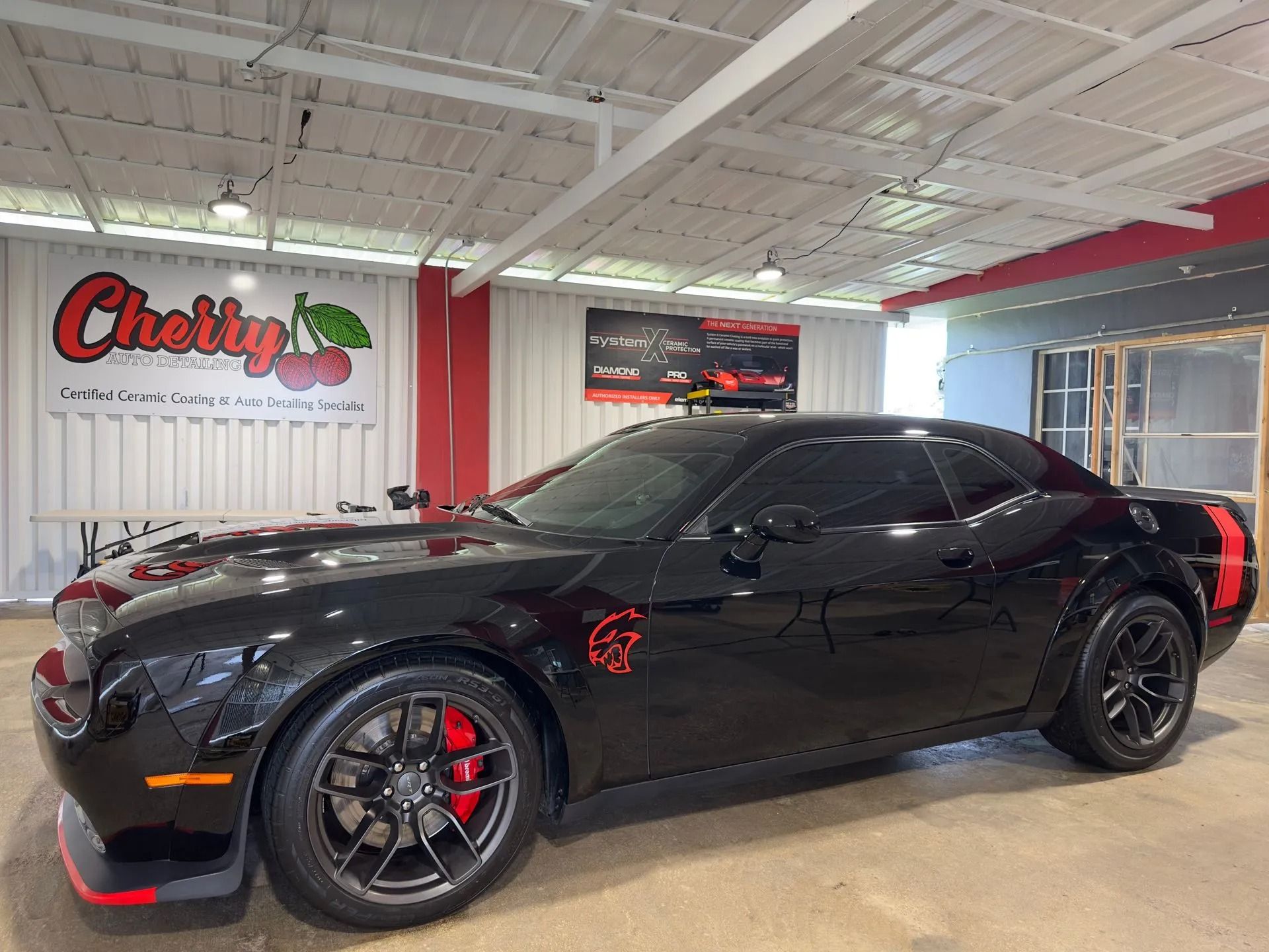 Black Dodge Challenger with red stripes and brake calipers parked inside a garage with a Cherry car logo on the wall.