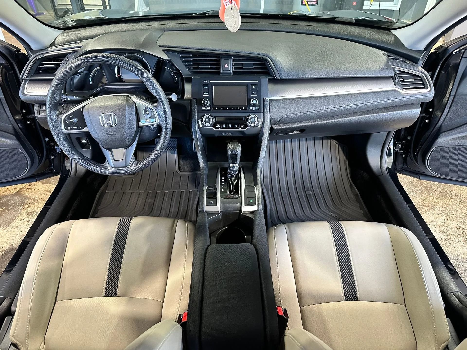 The interior of a vehicle featuring a grey dashboard, light-colored seats with dark accents, and black rubber floor mats.