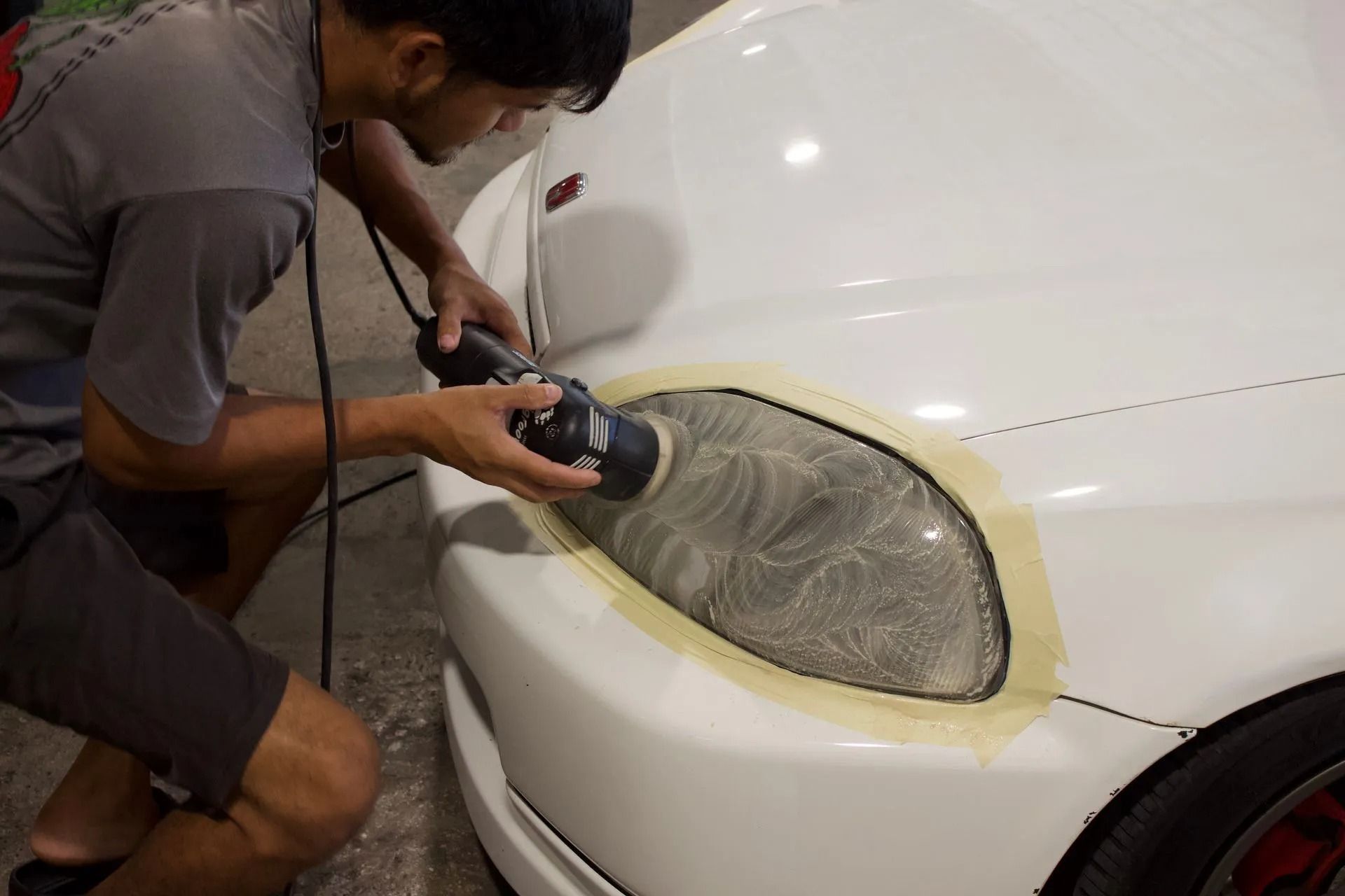 A person uses a power polisher to restore a cloudy headlight on a white car, with masking tape protecting the paint.