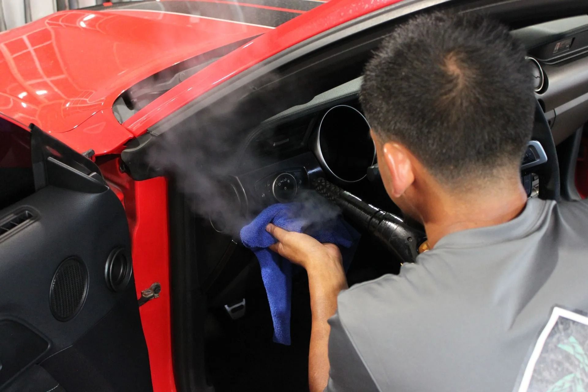 A technician uses a steam cleaner and a blue cloth to detail the dashboard of a red car.