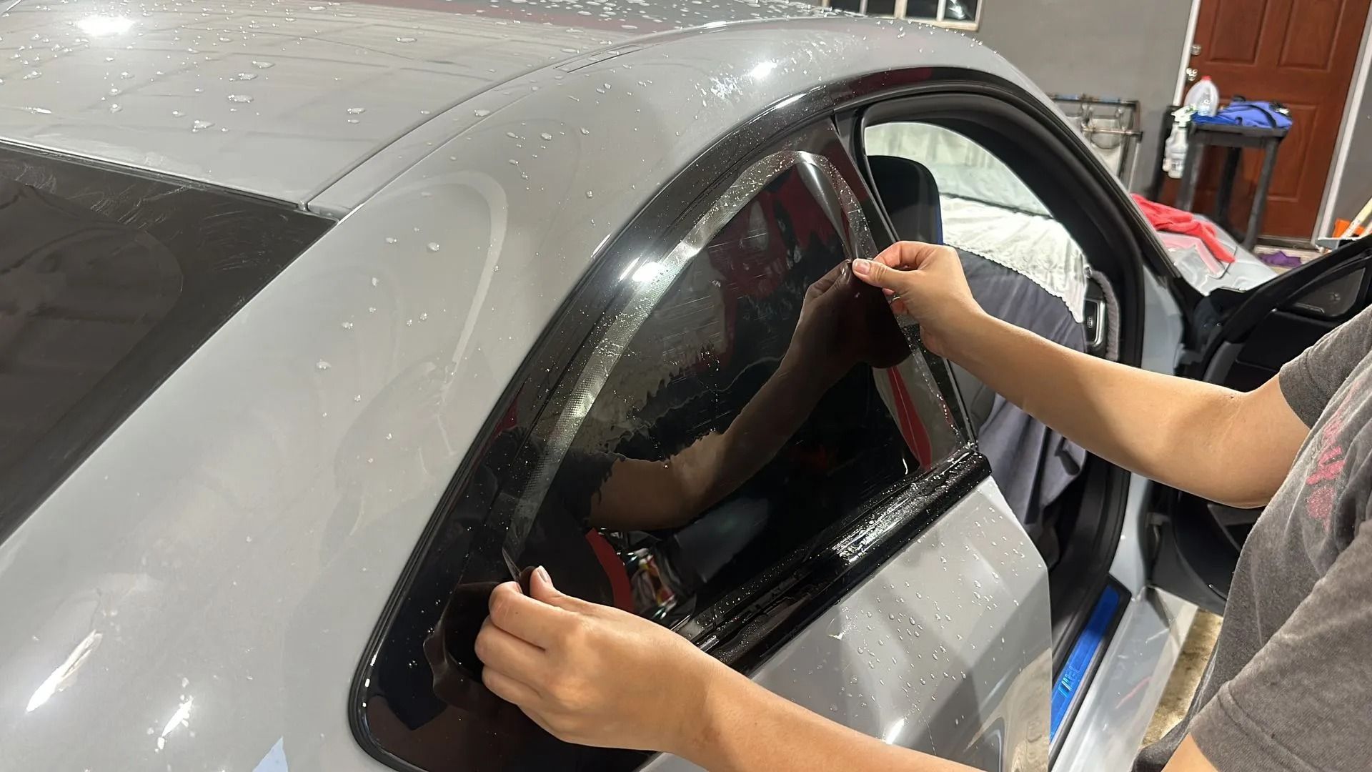 Hands applying a dark tint film to the rear passenger window of a grey car in a garage.