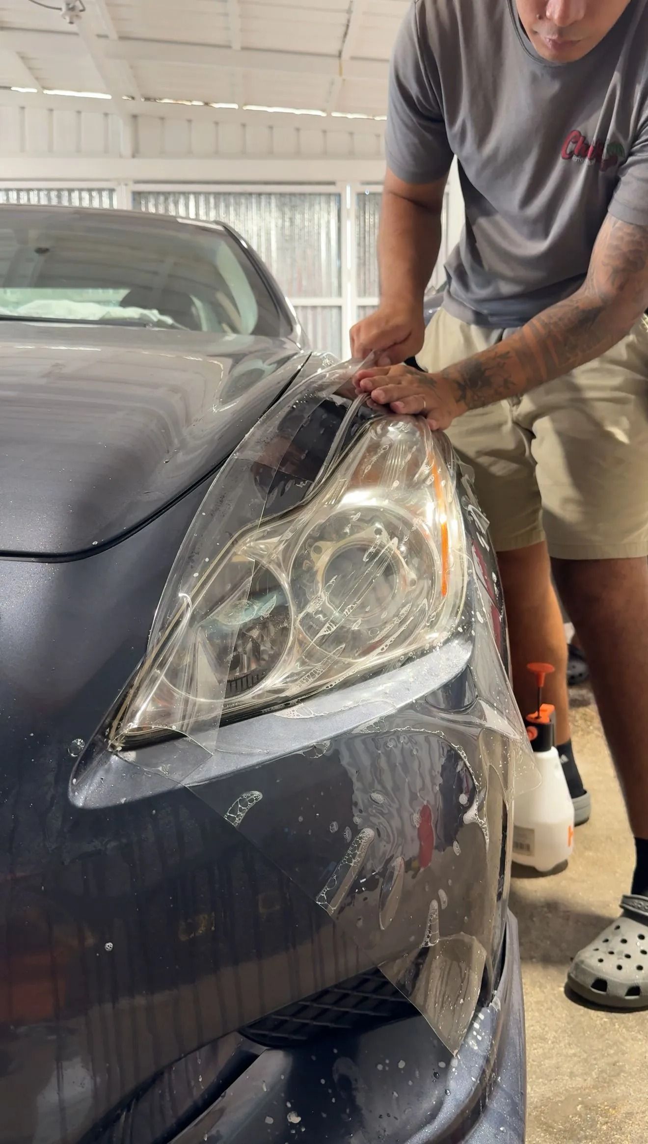 A person applying a clear protective film onto a vehicle's headlight in a garage.