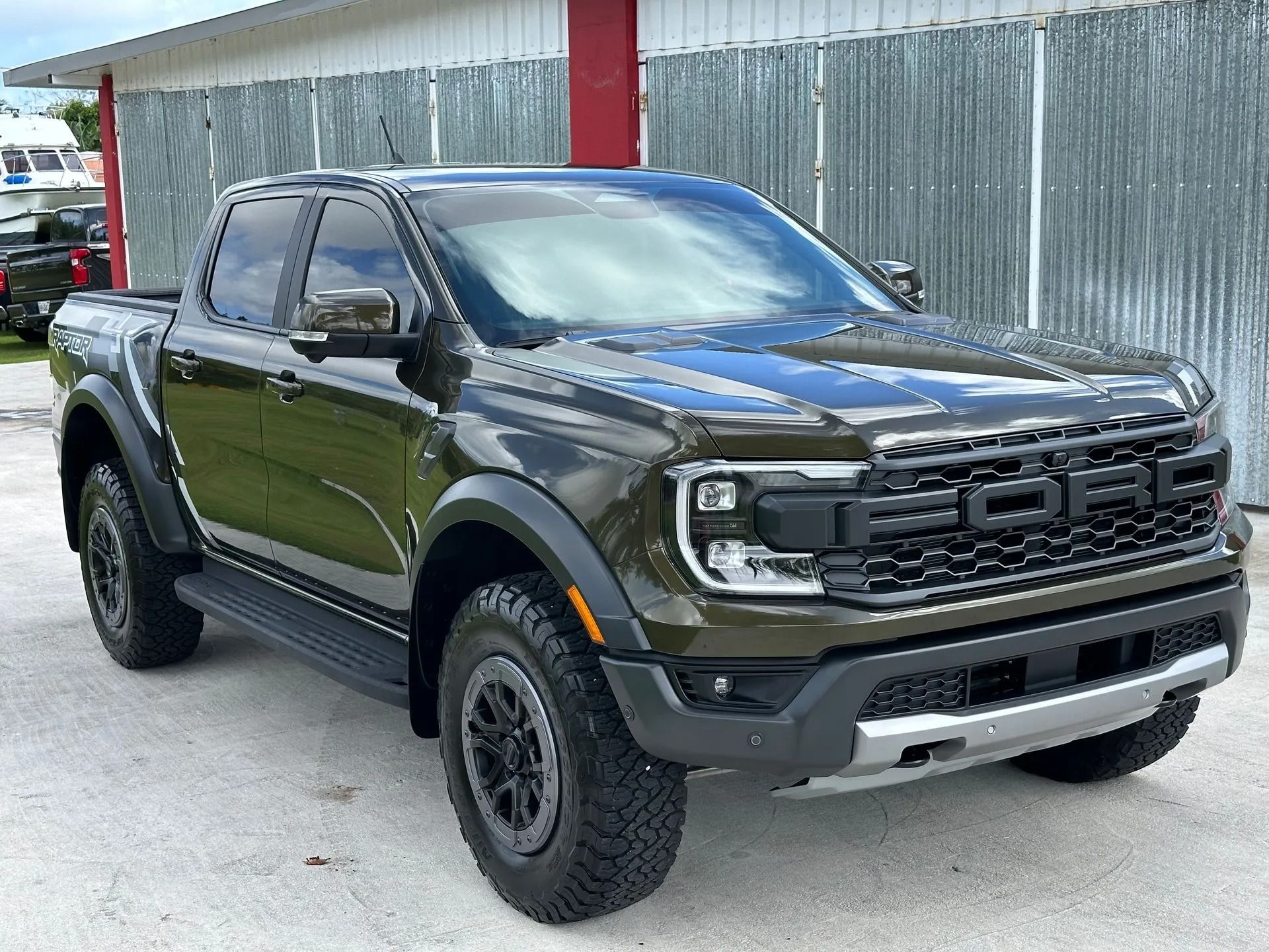 An olive green Ford Ranger Raptor pickup truck parked in front of a corrugated metal wall.