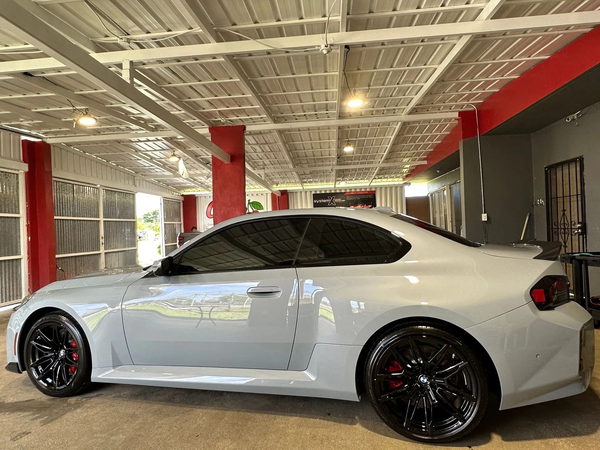 A light grey BMW M2 coupe parked in a well-lit, industrial-style garage with red columns and a white ceiling structure.