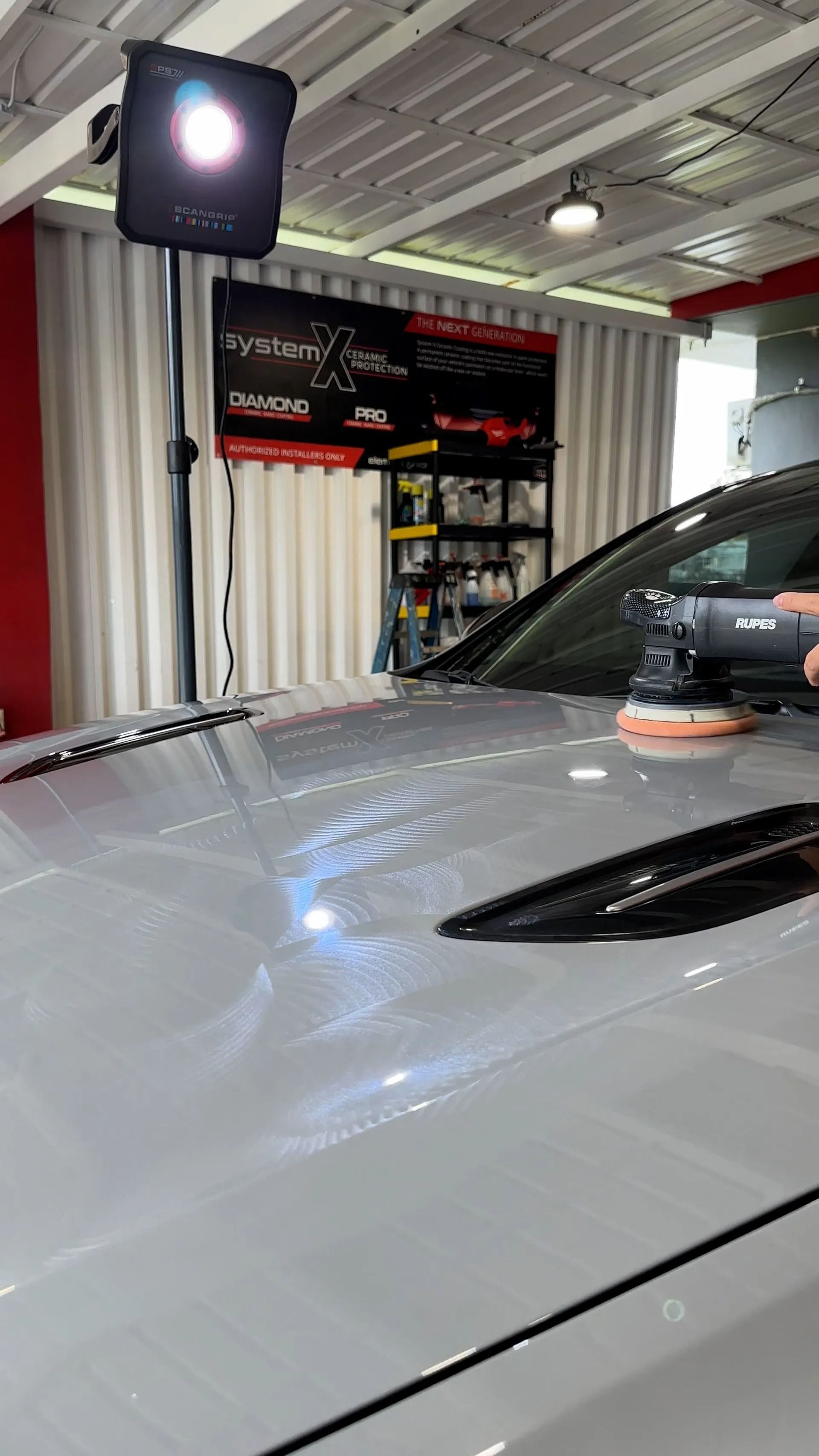 A detailer uses an orbital polisher on the hood of a silver car under bright garage lights.
