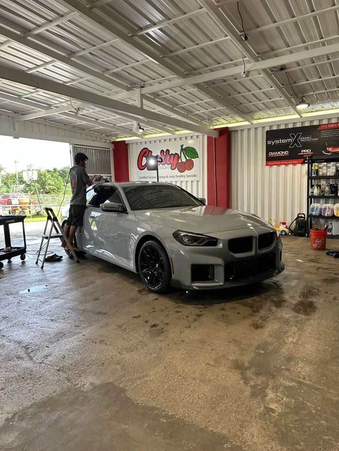 A technician works on a grey sports car inside an automotive detailing shop with a logo on the back wall.