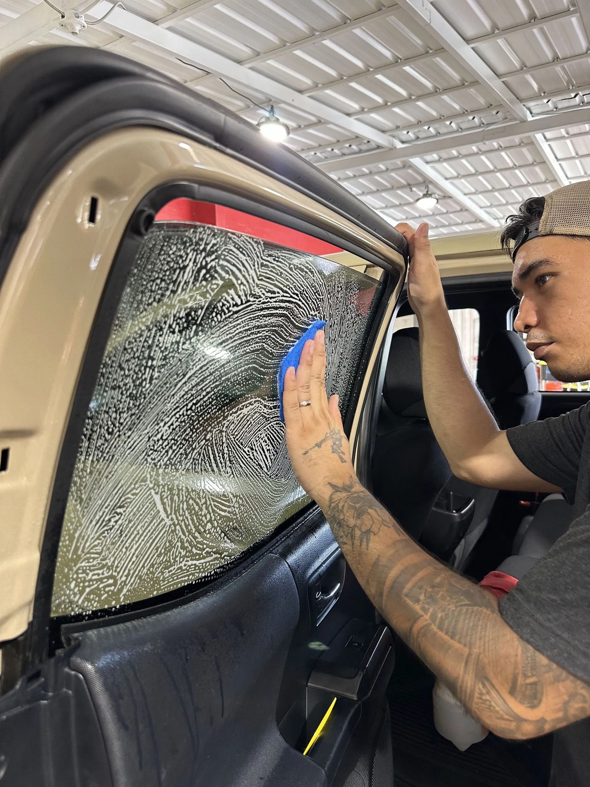 A technician applies window tint film to a vehicle door window using a blue squeegee in a brightly lit garage.