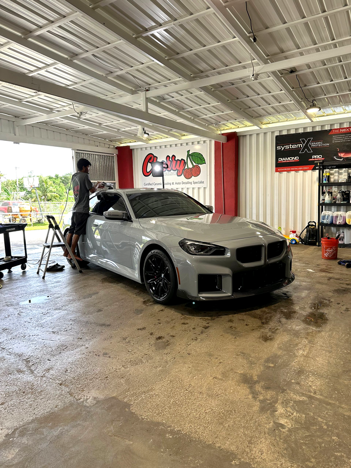 A person in a garage applies a wrap or coating to a gray BMW sports car.