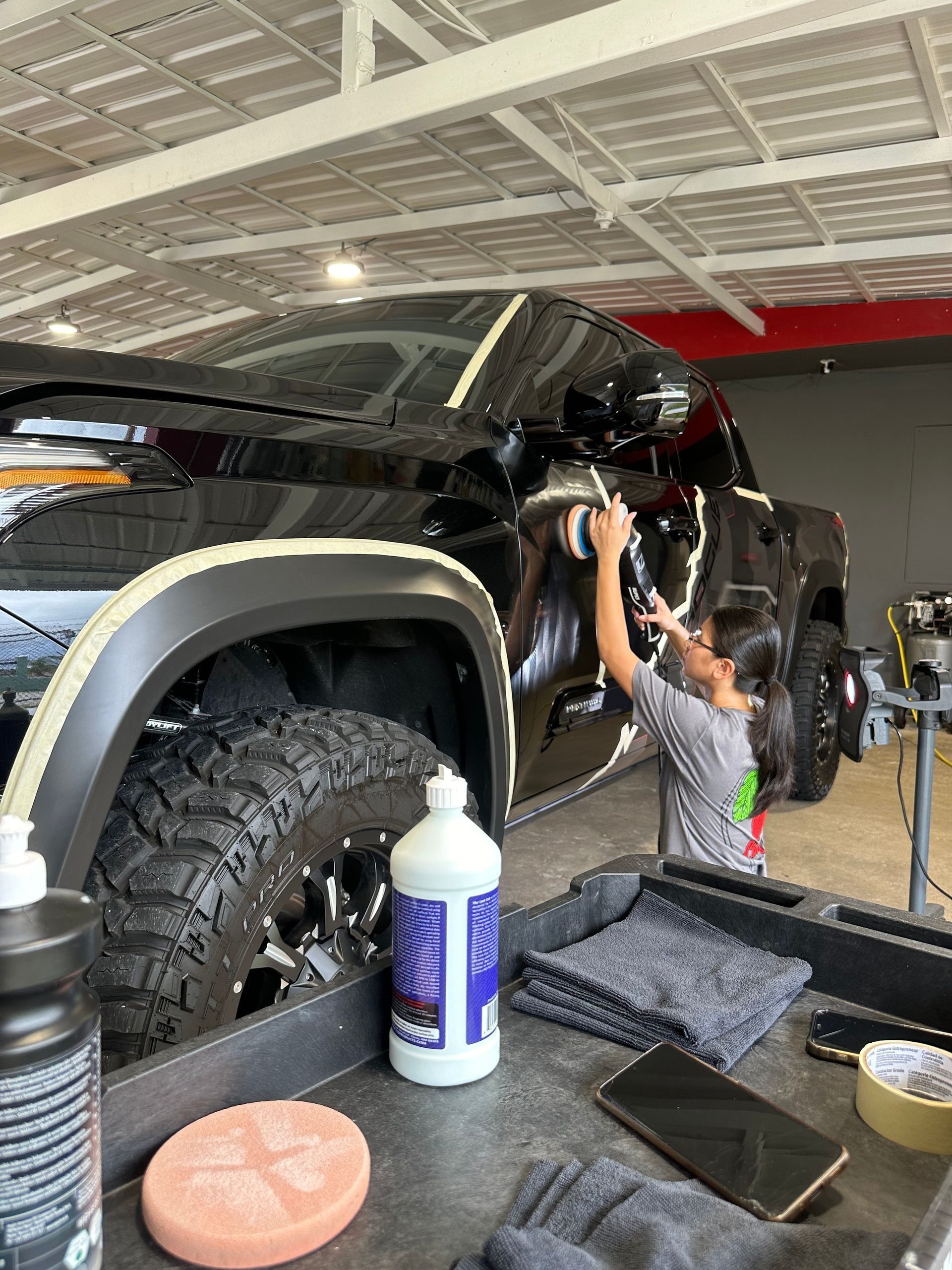 A person in a gray shirt uses a polisher on the side of a black truck inside a garage workspace.