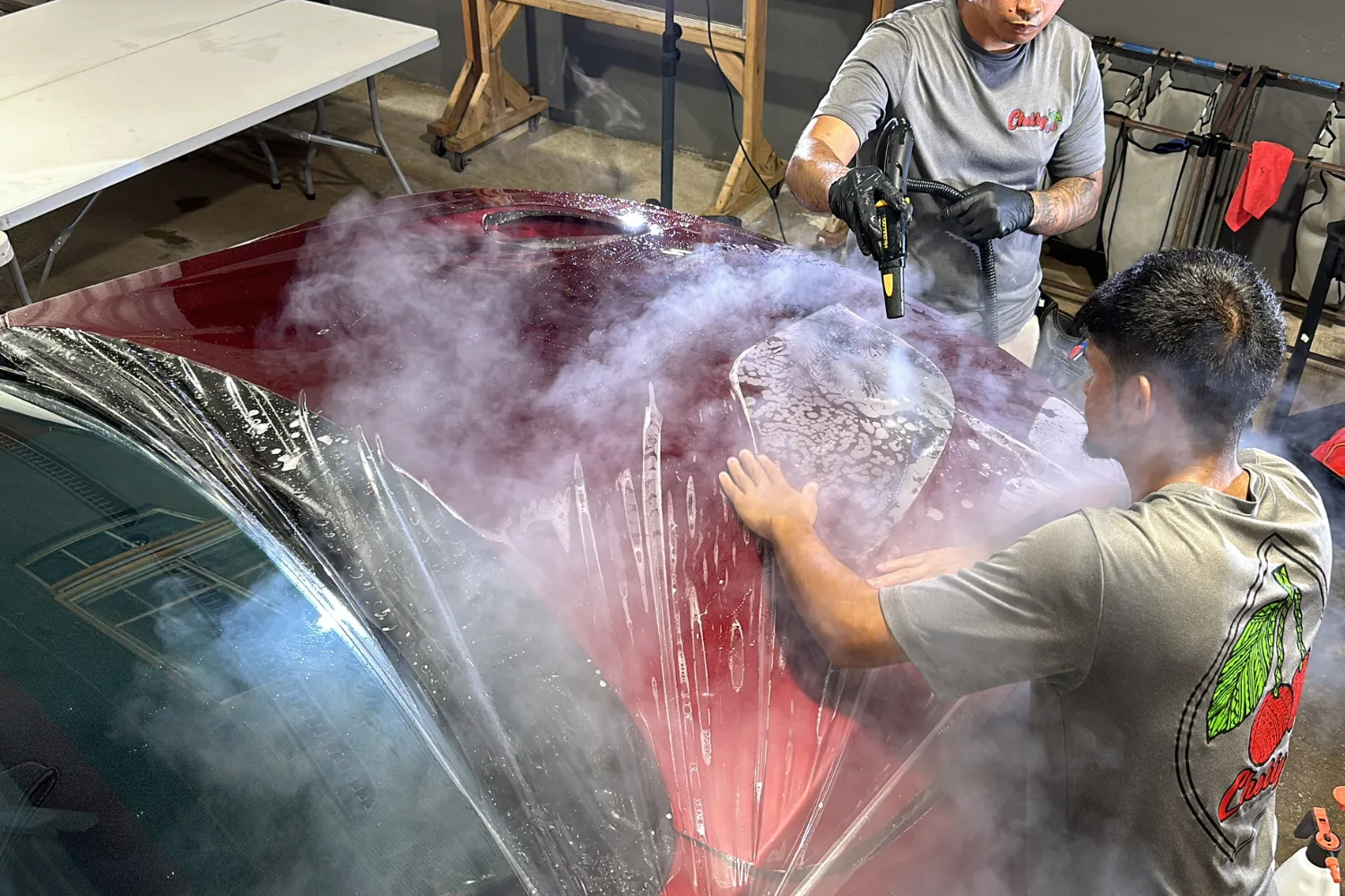Two technicians use steam and tools to apply a clear protective film to the hood of a dark red vehicle.