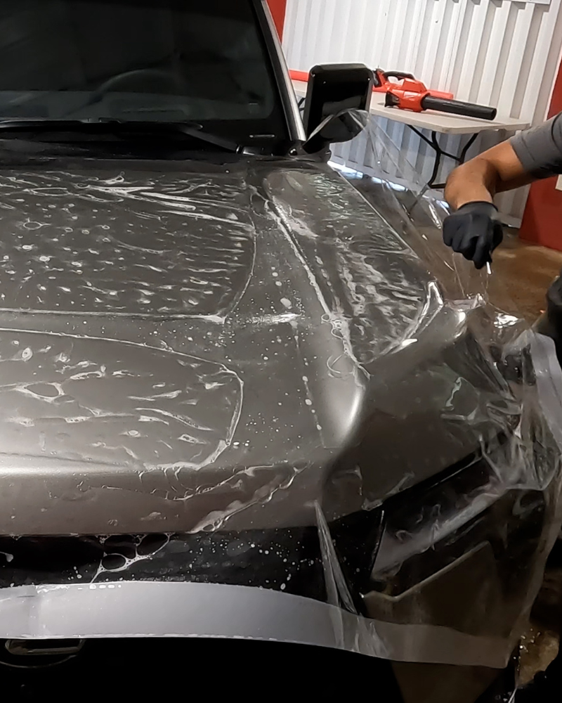 A technician in black gloves applies a clear protective film to the hood of a silver car.