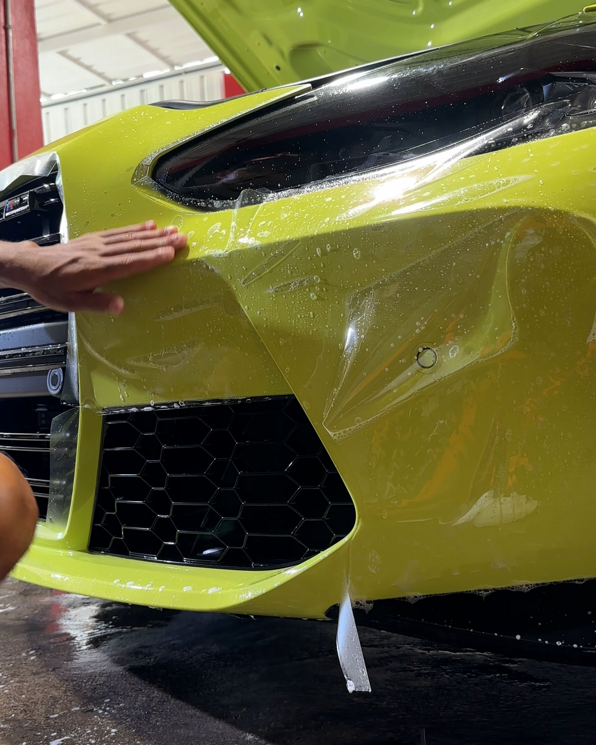 A hand applies a protective clear film to the lime green bumper of a car in a workshop.