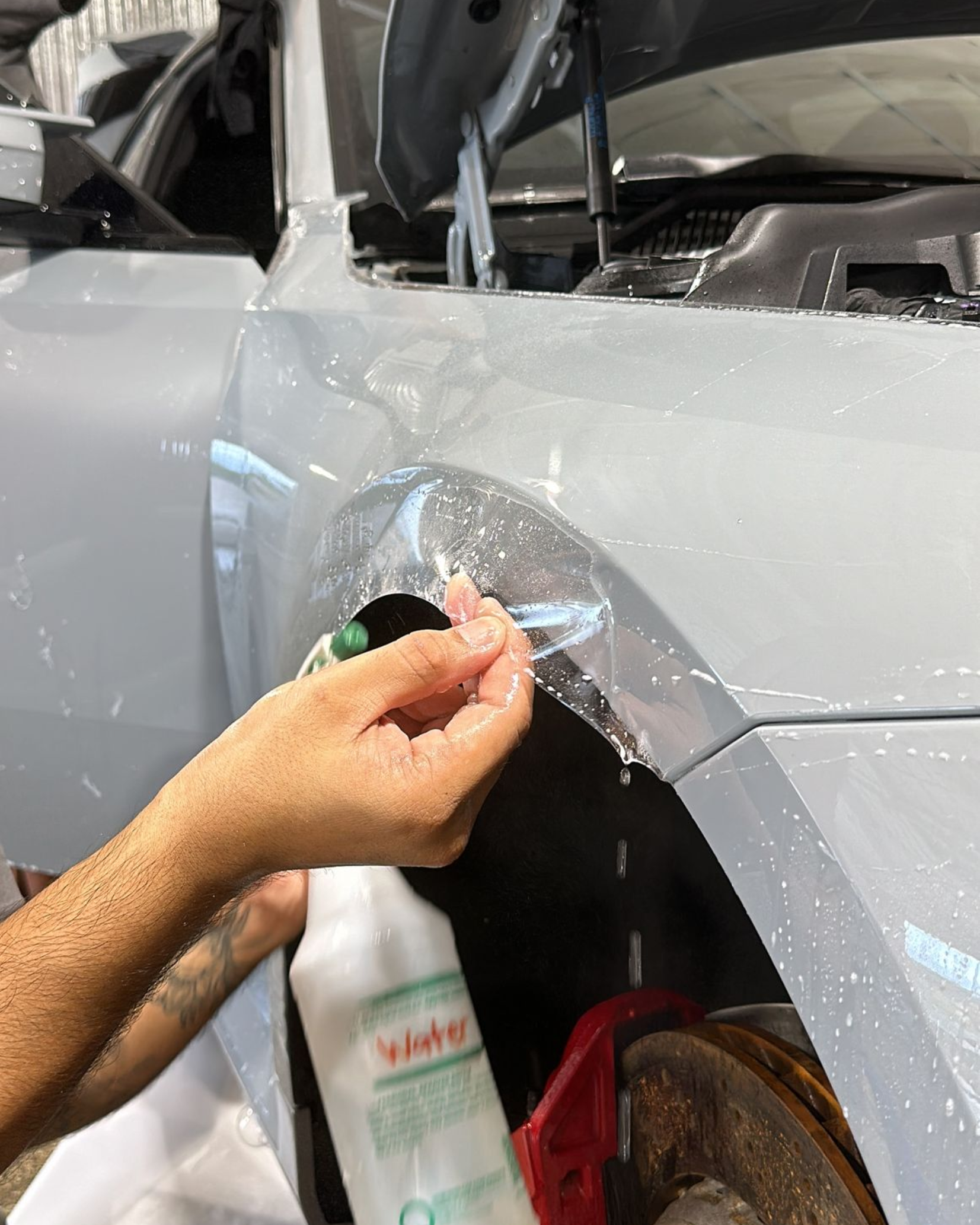 A person sprays solution on a clear paint protection film being applied to the wheel arch of a grey car.