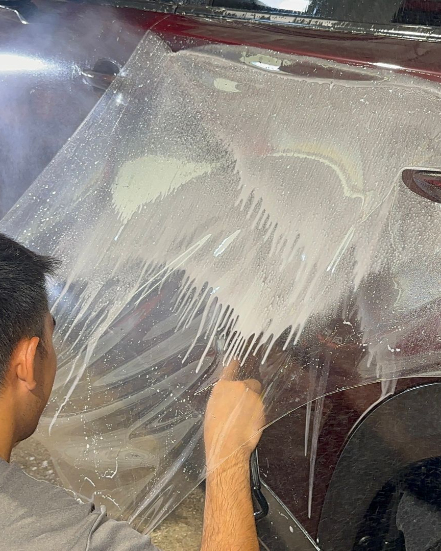 A person applying a clear protective film to the side of a red vehicle using a squeegee and soapy liquid.