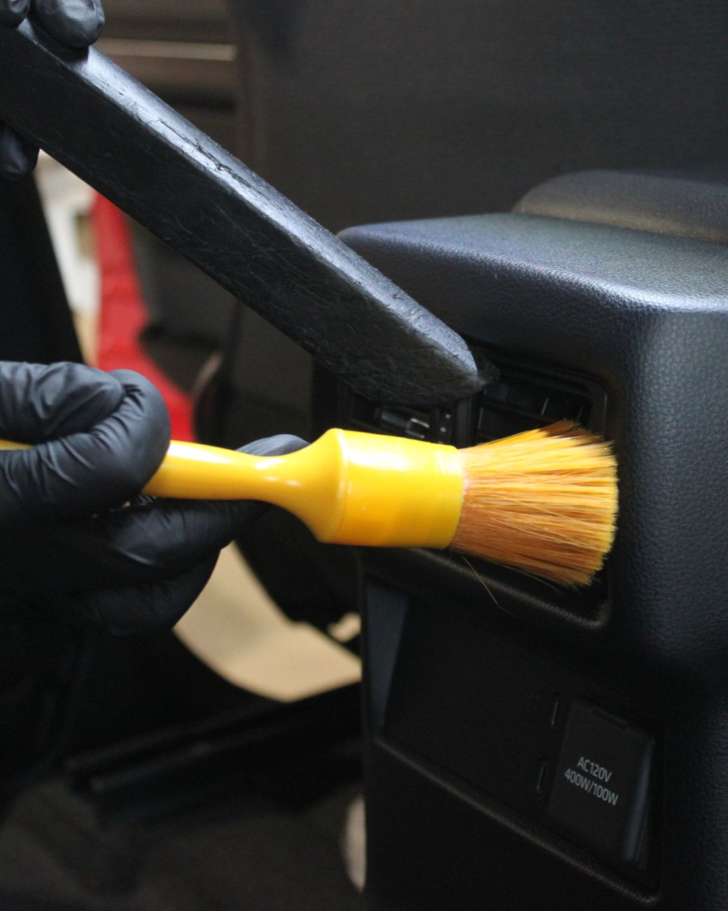 Gloved hands use a yellow detailing brush to clean dust out of a car's air vent while a vacuum nozzle sits nearby.